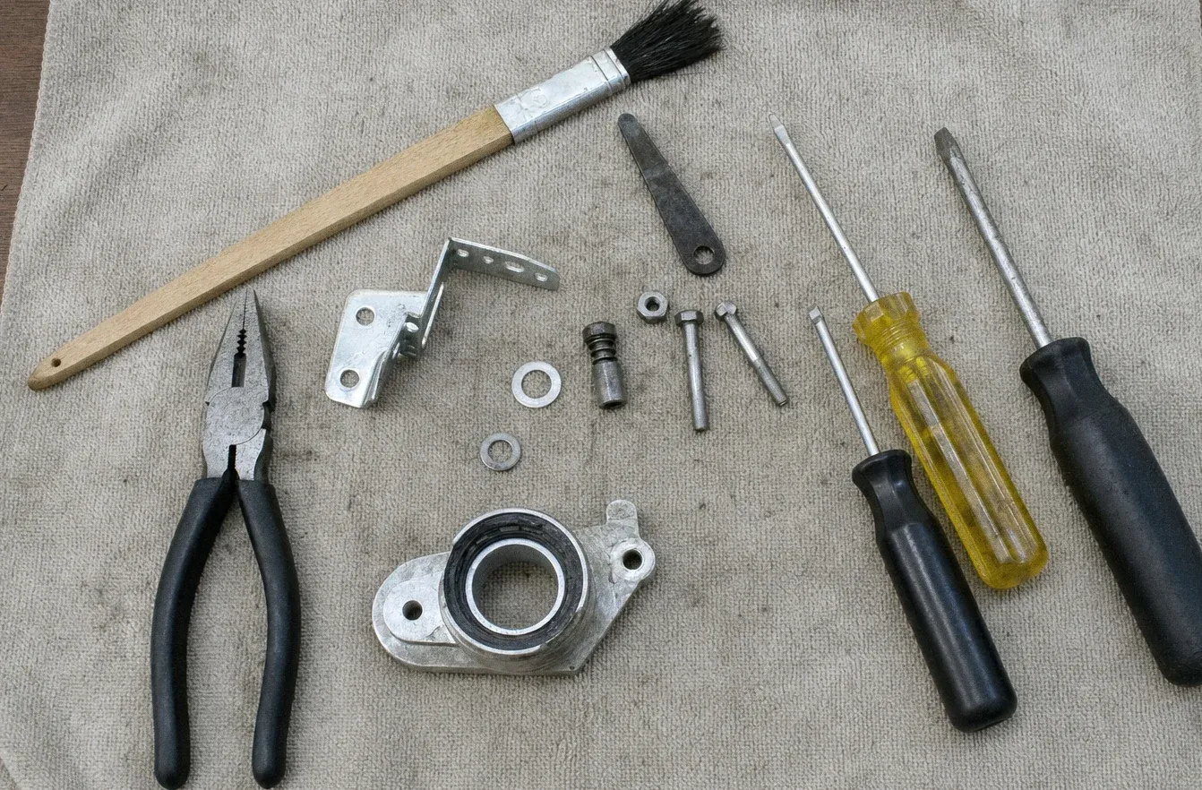Long-handled brush, needle-nose pliers, a hinge bracket, a burner base plate, spacers and washers on a cloth